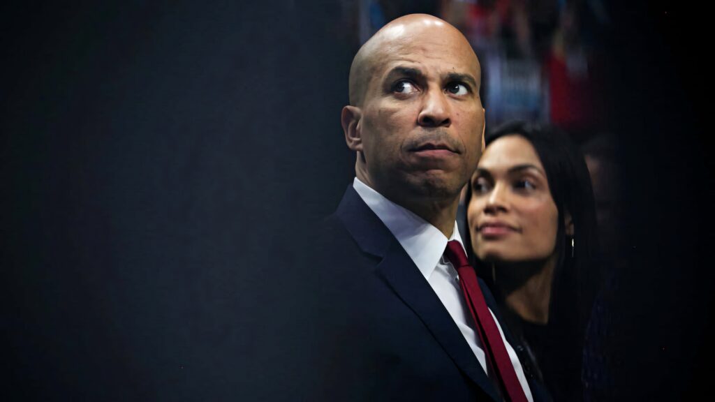 Cory Booker in formal suit with serious expression and supportive companion dark background wallpaper