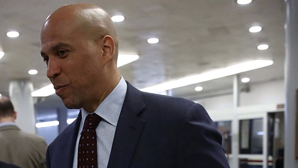 Side view of Cory Booker in navy suit with blurred modern indoor background wallpaper