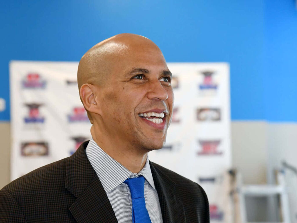 Cory Booker smiling warmly in business attire with vibrant blue background and event signage Wallpaper
