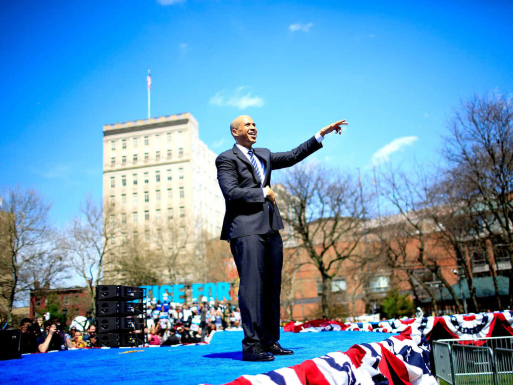 Cory Booker speaking outdoors with patriotic decorations and urban skyline at public event Wallpaper