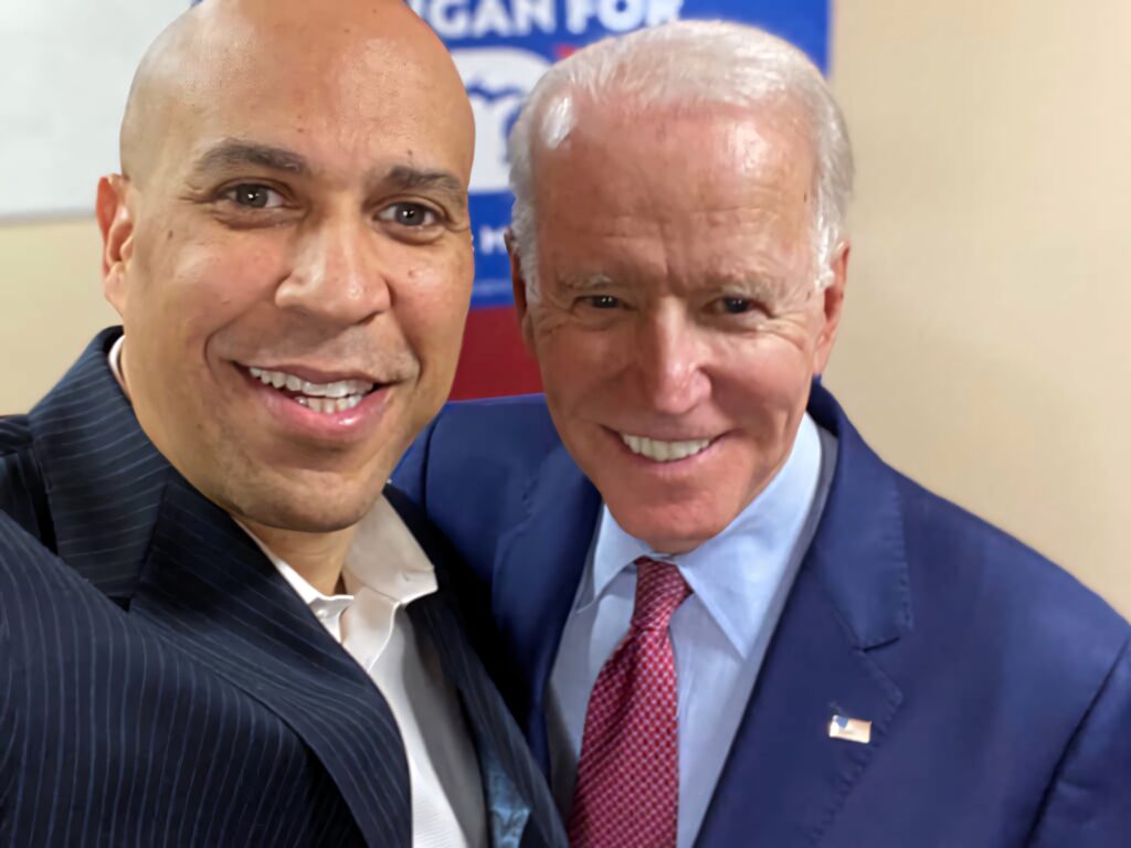Cory Booker and President Biden smiling in suits with blurred campaign sign background wallpaper