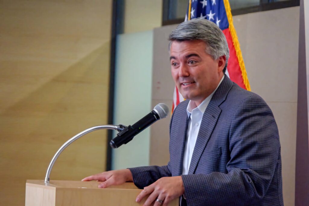 Cory Gardner speaking confidently at podium with American flag and warm lighting background Wallpaper