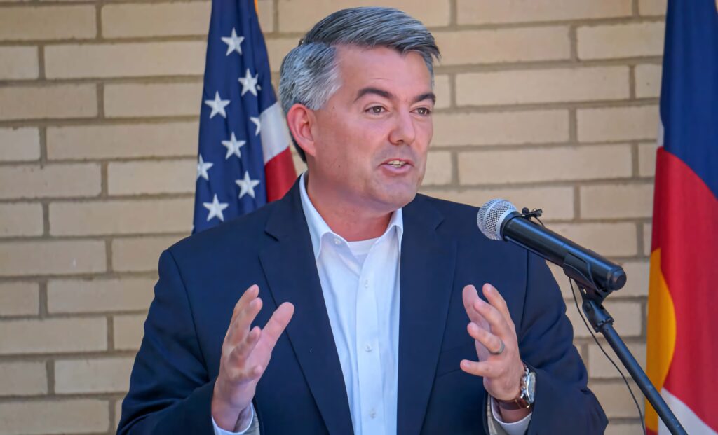 Cory Gardner speaking at podium with American and state flags in navy suit white shirt brick wall Wallpaper