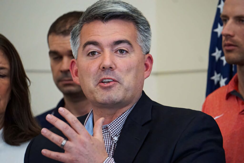 Cory Gardner speaking at event in dark blazer checkered shirt with American flag background wallpaper