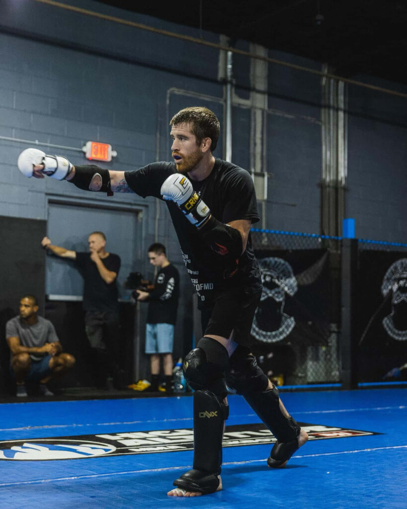 Cory Sandhagen mid-action throwing punches in black gear at modern MMA gym with blue mats Wallpaper