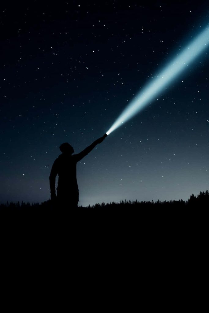 Person pointing a bright flashlight beam into a starry night sky with cosmic background wallpaper
