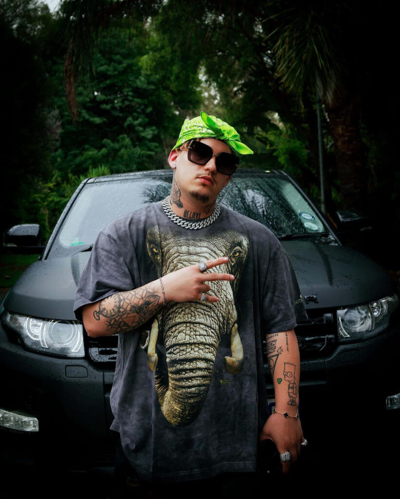 Costa Titch in elephant print shirt and chains by dark SUV with moody urban greenery background Wallpaper