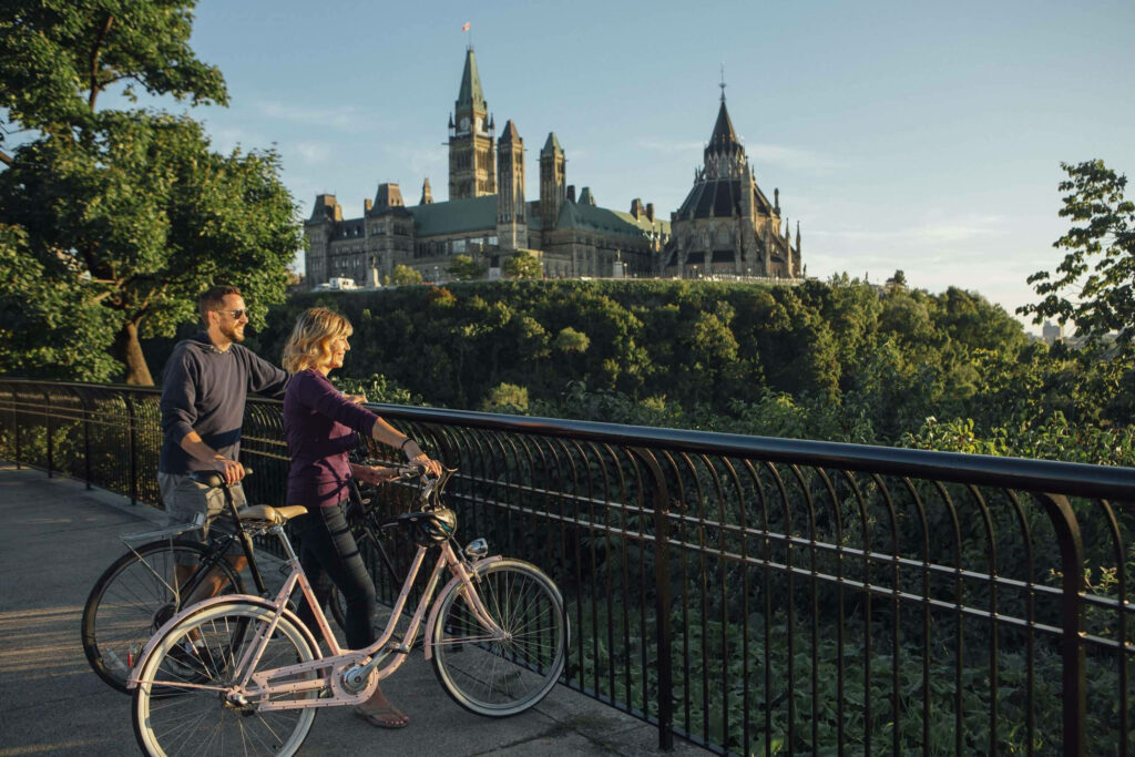 Couple cycling along Rideau Canal with Ottawa Parliament buildings and lush greenery in sunlight Wallpaper