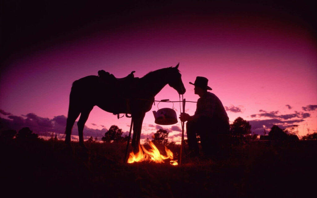 Cowboy tending campfire next to horse at vibrant purple pink sunset Western ranch silhouette Wallpaper