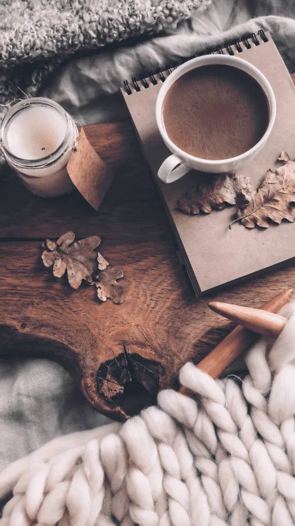Cozy autumn scene with warm coffee cup knitting yarn spiral notebook dried leaves candle Wallpaper