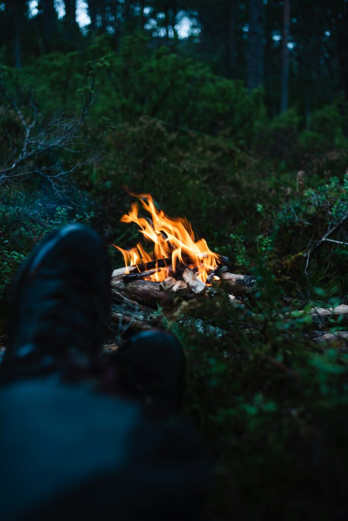 Cozy bonfire glowing in green forest clearing with relaxed legs camping nature wallpaper background