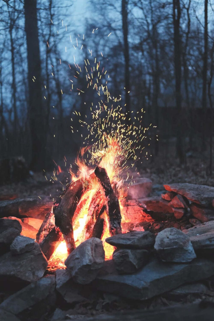 Warm campfire glowing with sparks surrounded by stones and bare trees under starlit sky Wallpaper