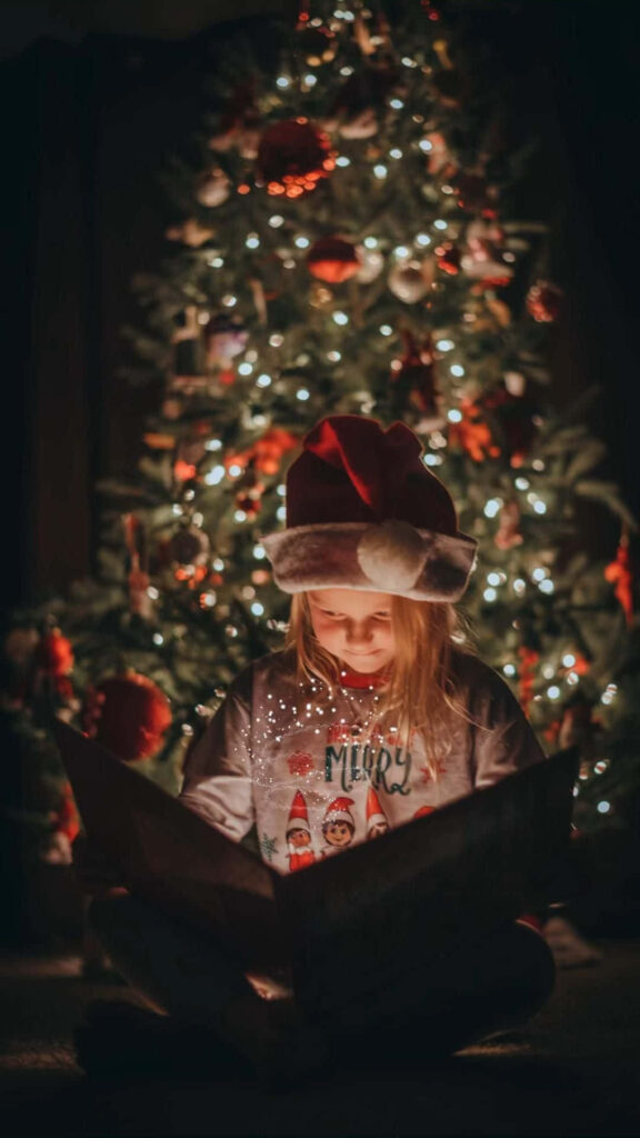 Cozy Holiday Magic: Girl in Santa Hat Reads by Christmas Tree Wallpaper