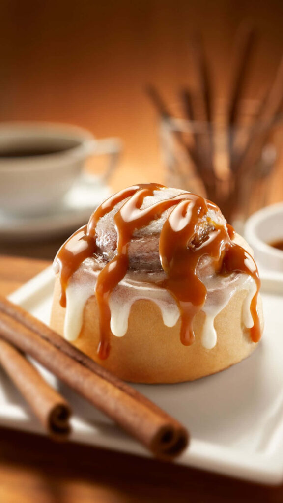 Freshly baked cinnamon roll with creamy icing and caramel sauce on white plate with coffee Wallpaper