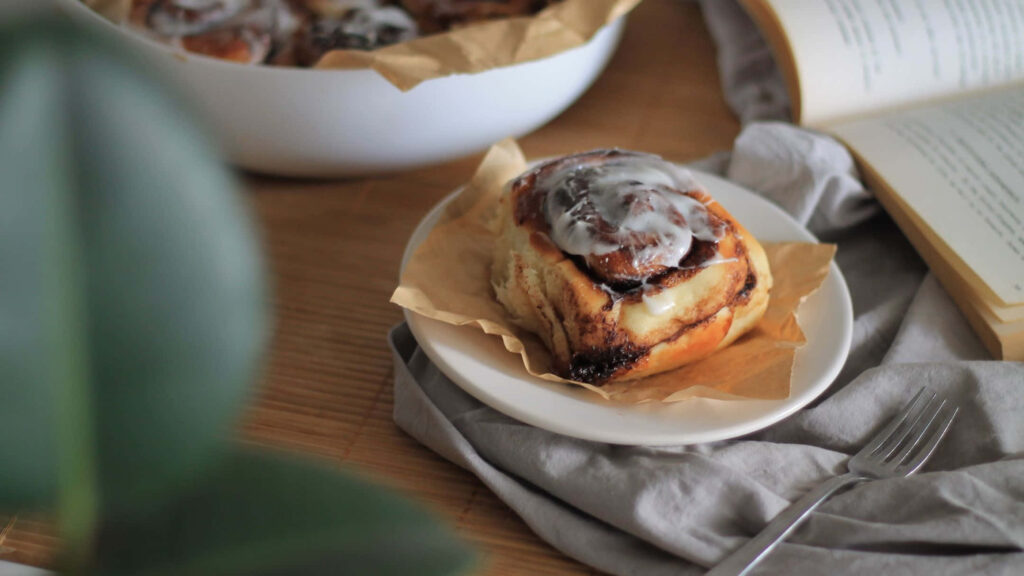Fresh cinnamon roll with glossy icing on white plate cozy wooden table open book greenery Wallpaper
