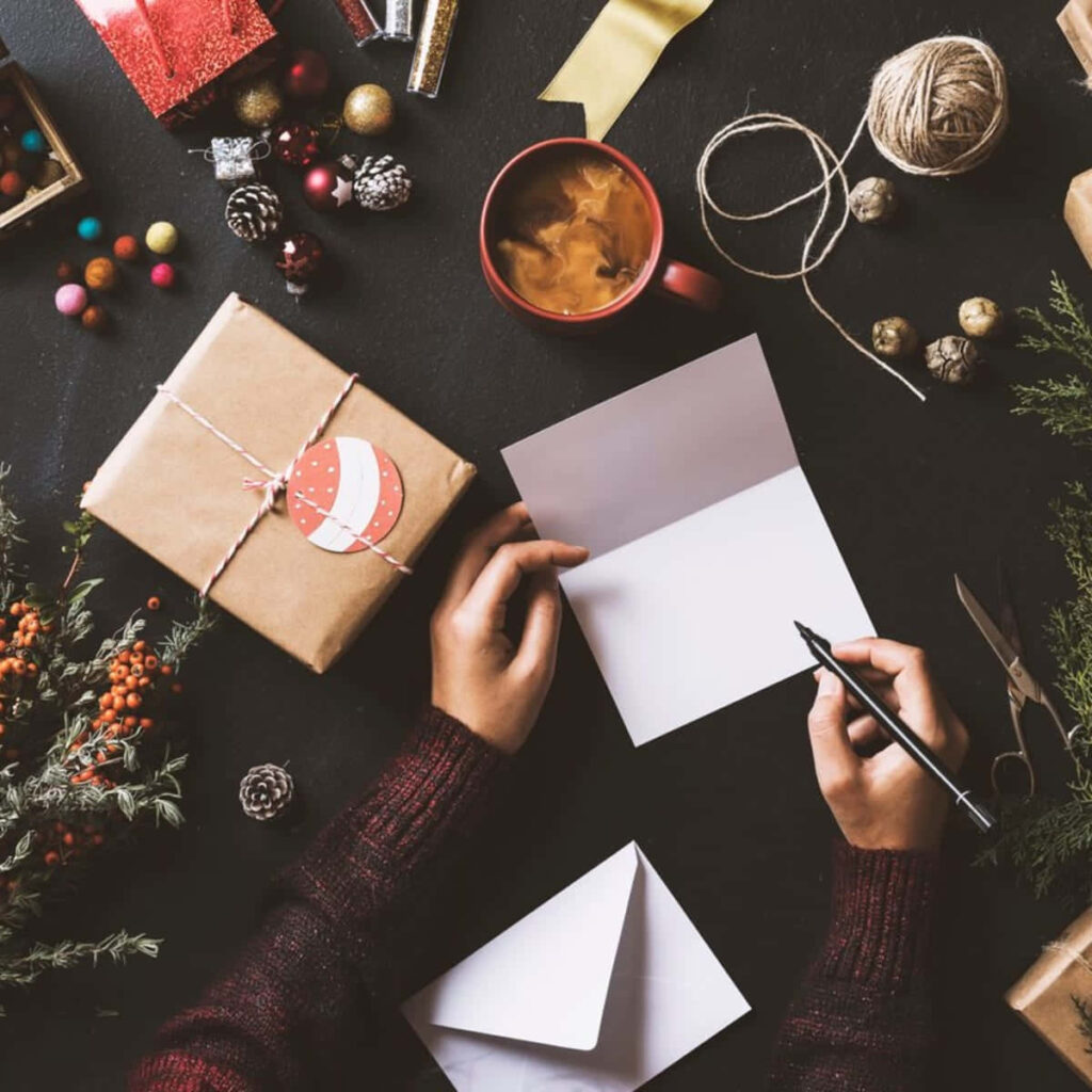 Cozy Holiday Scene: Writing Cards Amidst Festive Gifts & Greenery Wallpaper