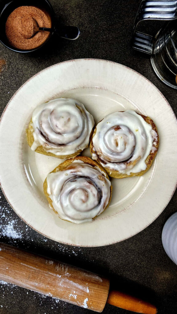 Three frosted cinnamon rolls on cream plate with baking tools warm cozy homemade vibe wallpaper