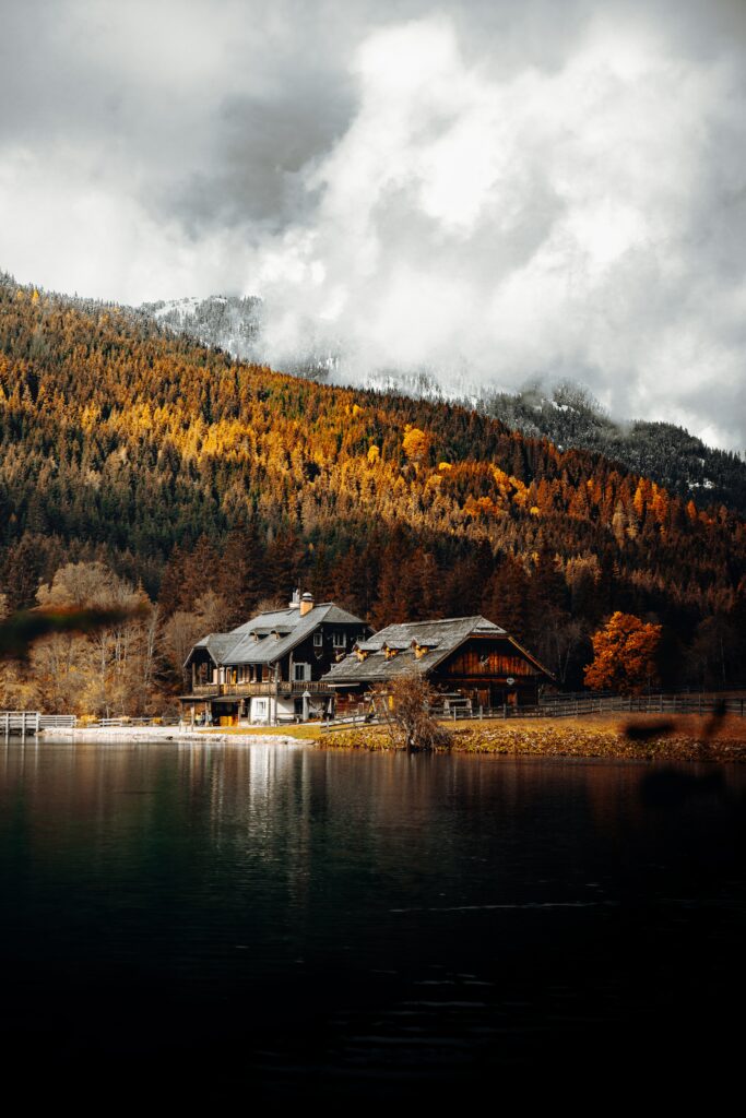 Cozy lakeside house surrounded by vibrant autumn forest reflections under cloudy sky wallpaper