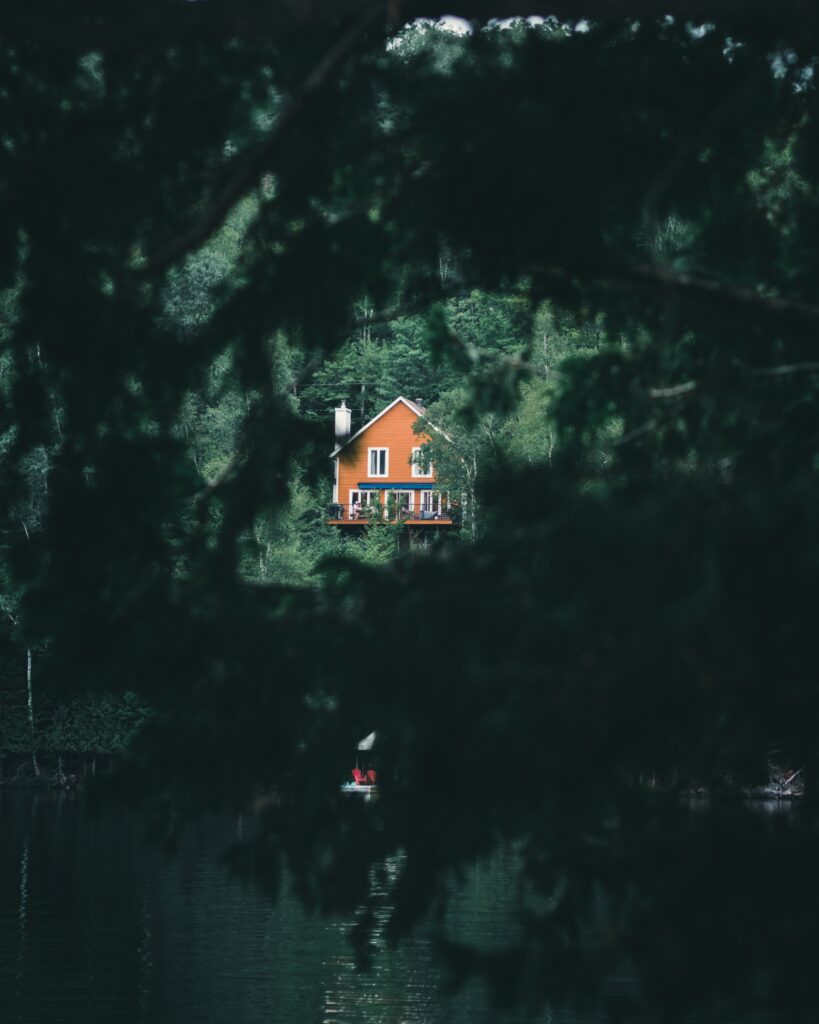 Cozy warm-toned house by peaceful lake surrounded by lush green forest reflected in calm water Wallpaper