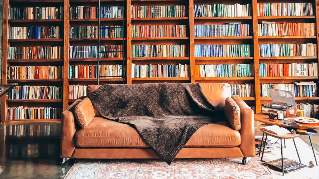 Cozy brown leather sofa with dark knit blanket by tall wooden library perfect Zoom background Wallpaper