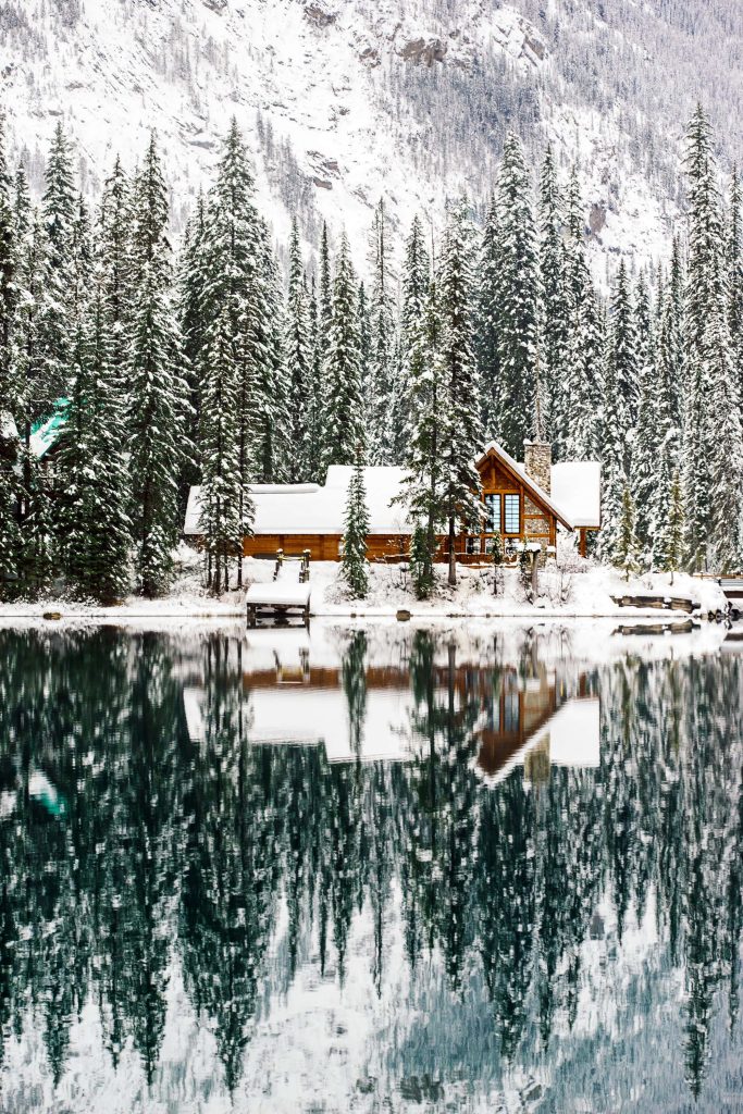 Cozy log cabin in snowy evergreen forest reflecting on tranquil lake with distant snowy peaks Wallpaper