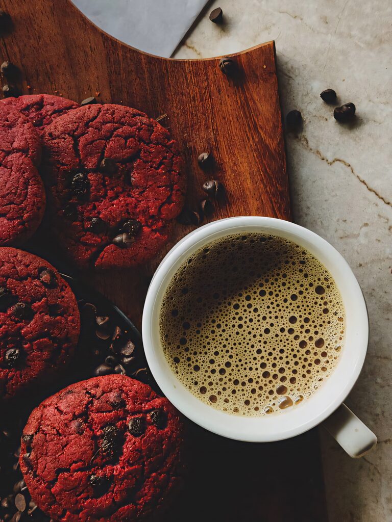 Red velvet chocolate chip cookies and steaming coffee on wooden board cozy café wallpaper background