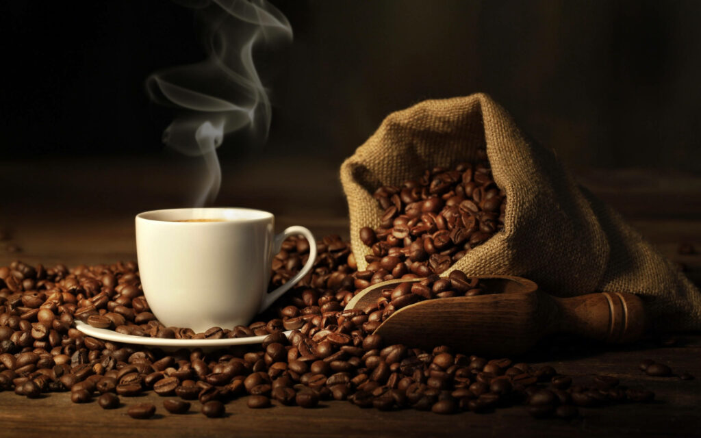 Steaming white coffee cup with burlap sack and wooden scoop surrounded by rich coffee beans Wallpaper