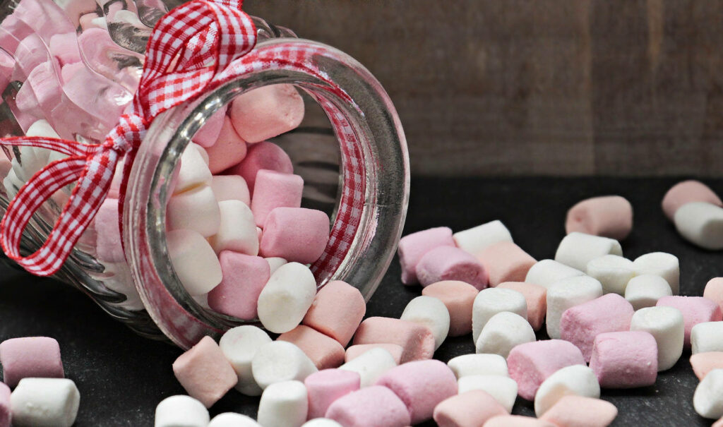 Glass jar with red gingham ribbon spilling pink and white marshmallows on dark rustic surface Wallpaper