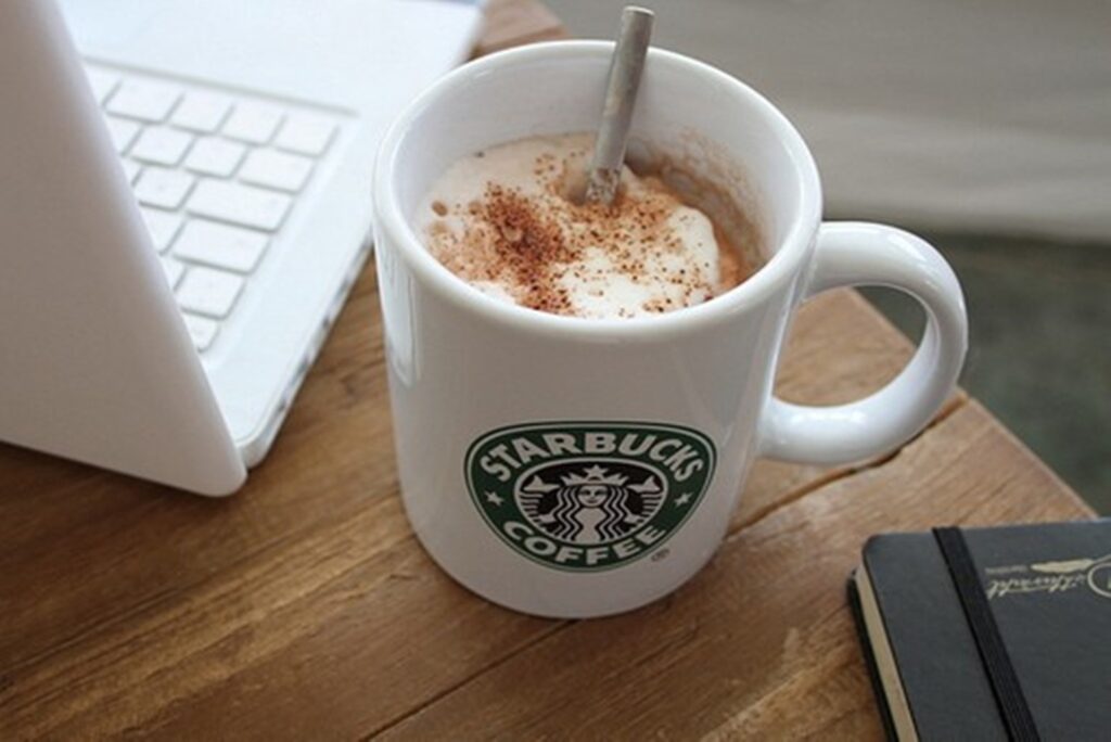 Cozy Starbucks Setup: Frothy Mug, Laptop & Notebook on Wooden Table Wallpaper