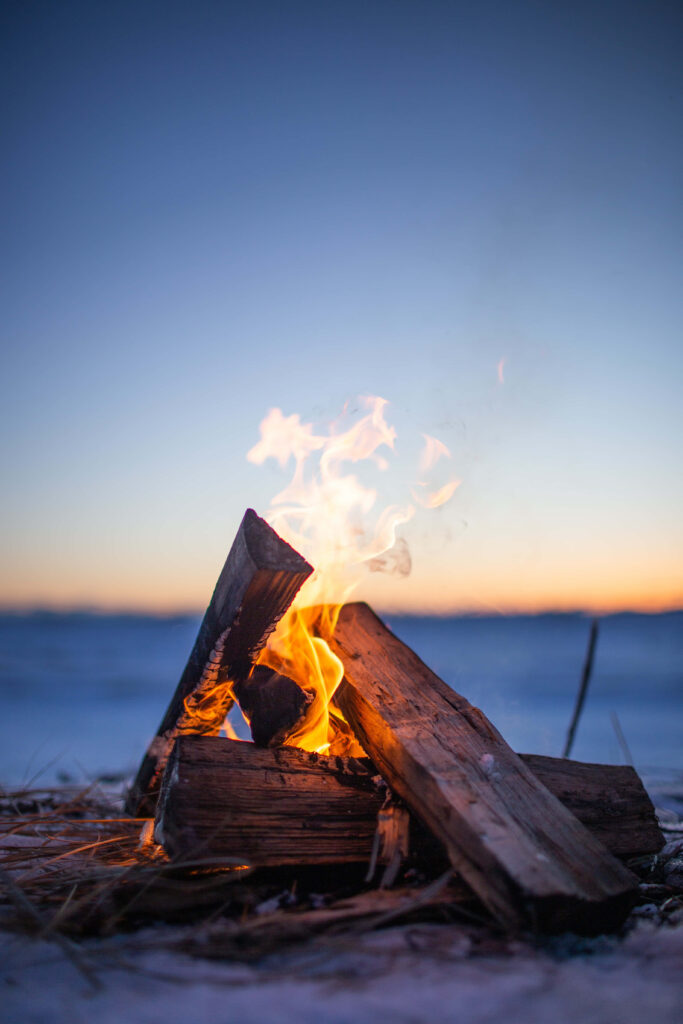 Cozy campfire glowing on snowy ground with wooden logs beneath vibrant winter sunset sky background Wallpaper