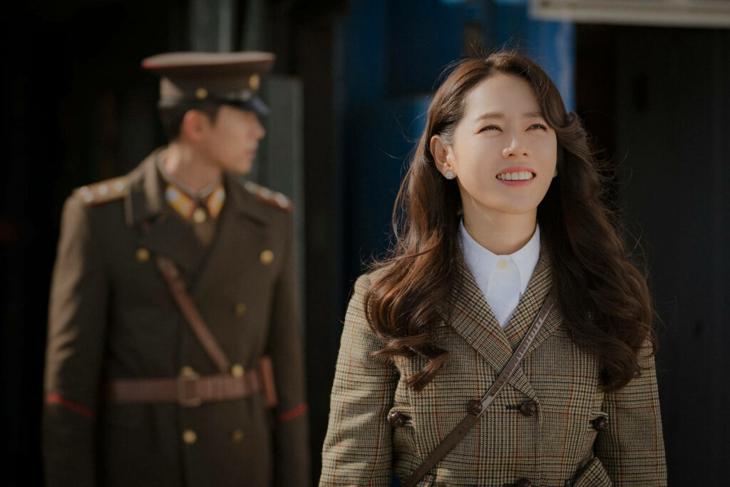 Yoon Se-ri smiling in checked coat with blurred Ri Jeong-hyeok military uniform Crash Landing On You Wallpaper
