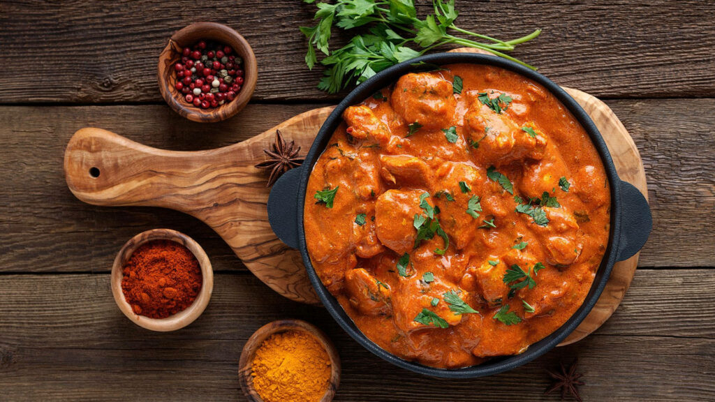 Butter chicken in rustic iron pan garnished with cilantro surrounded by spices on wood background Wallpaper