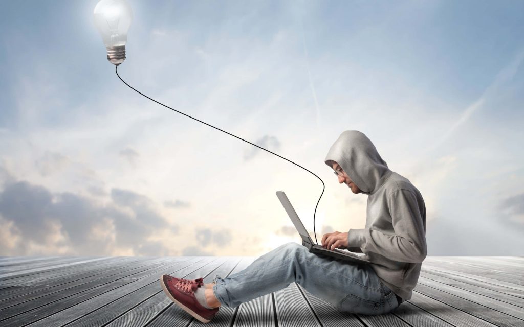 Man in grey hoodie typing on laptop with glowing lightbulb floating in cloudy sky background Wallpaper