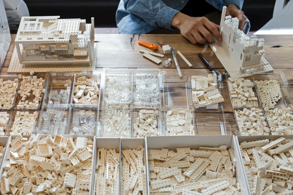 Person assembling detailed white Lego structures at wooden table with organized Lego trays Wallpaper