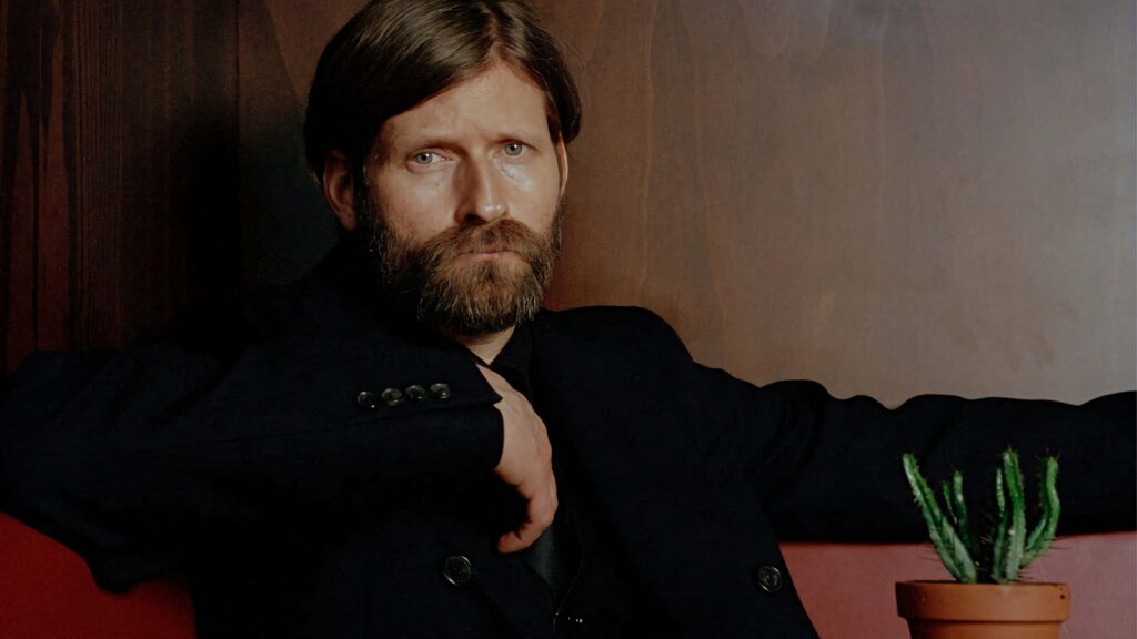 Crispin Glover in dark suit seated by wooden backdrop with potted cactus minimalist wallpaper