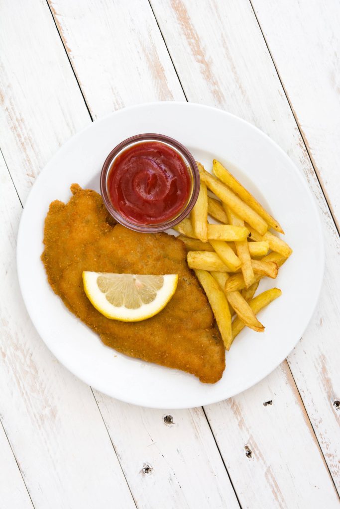 Crispy schnitzel with golden fries lemon and ketchup on white plate rustic wooden background Wallpaper