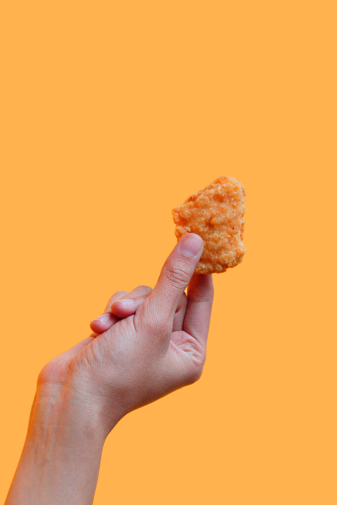 Close-up of hand holding crispy fried chicken against vibrant orange bold minimalist wallpaper background