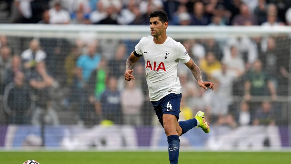 Cristian Romero in Tottenham Hotspur kit sprinting on pitch with stadium lights and blurred crowd background Wallpaper