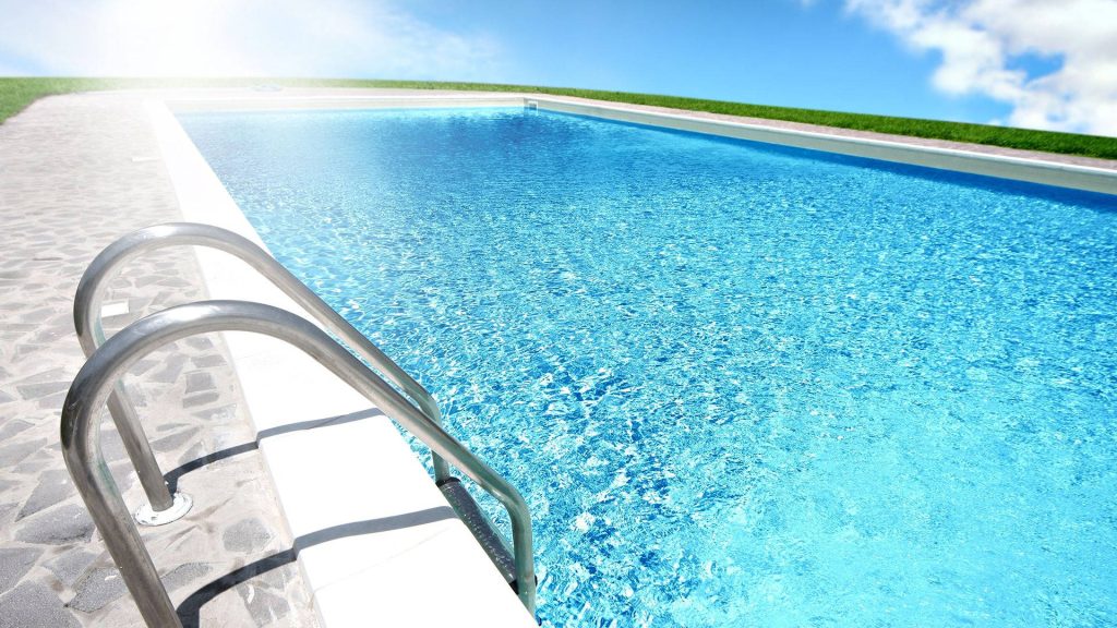 Polished stainless steel handrail into clear blue pool water with lush grass and stone deck Wallpaper