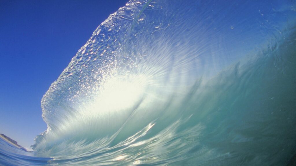 Crystal clear ocean wave curling under bright blue sky with sunlight shimmering sea splash wallpaper