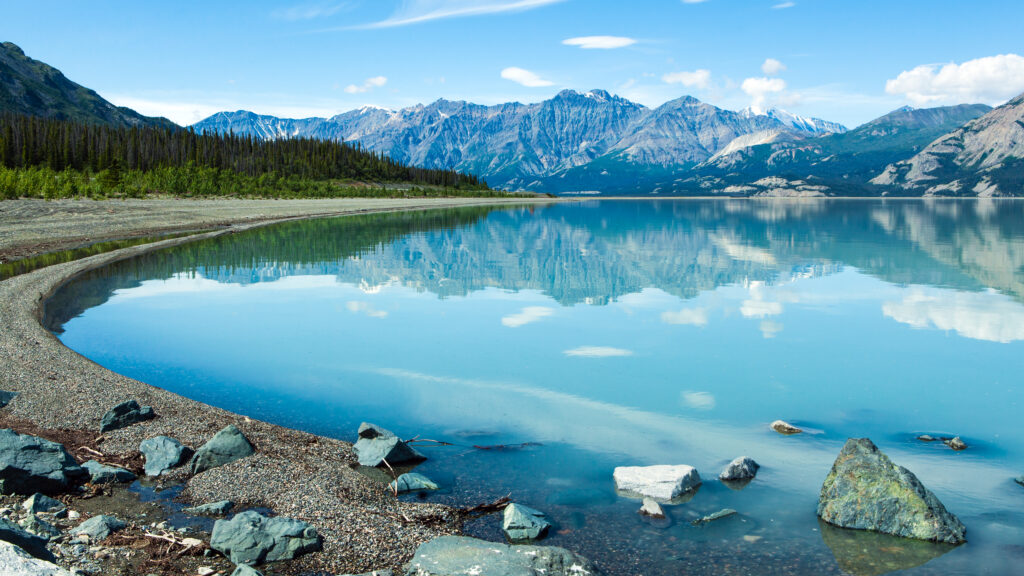 Crystal-clear lake reflecting blue mountains bright sky rocky shoreline and dense green forest Wallpaper