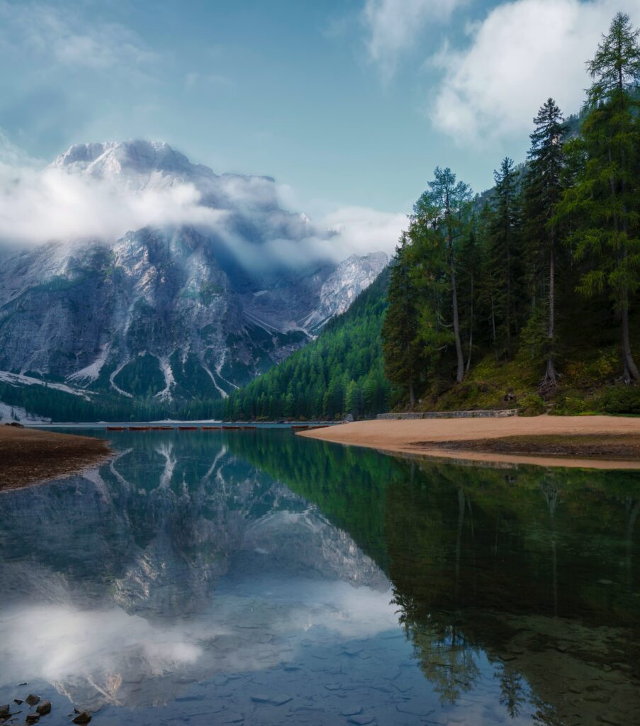 Serene lake reflecting pine trees dense forest and snow-capped mountains under partly cloudy sky Wallpaper