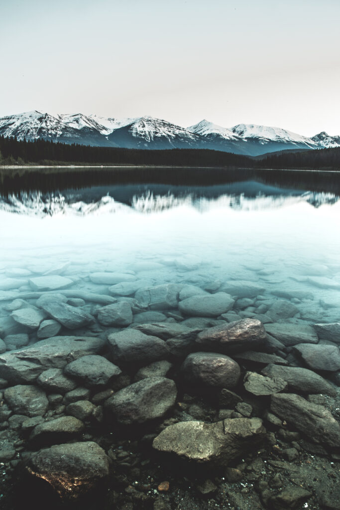 Crystal-clear lake reflecting snow-capped mountains with smooth rocks and a pale sky wallpaper