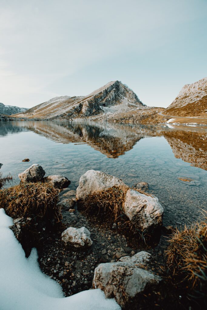 Crystal-clear lake reflecting majestic mountains with rocky shore under soft blue sky wallpaper