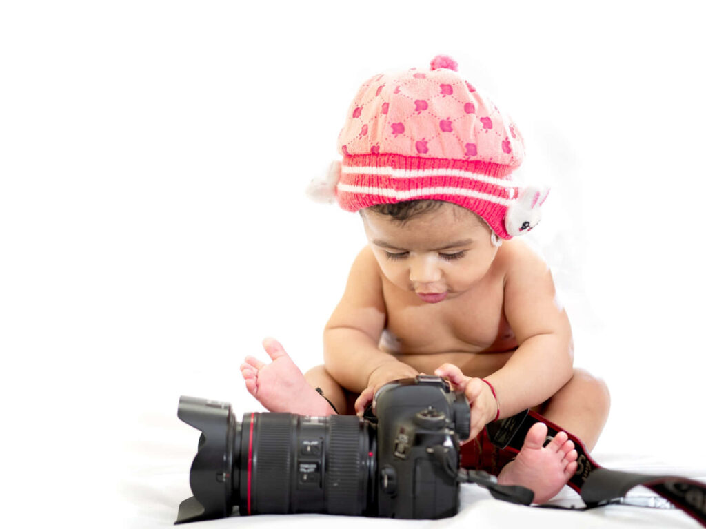 Joyful child in pink heart knit hat exploring professional camera on soft white background Wallpaper