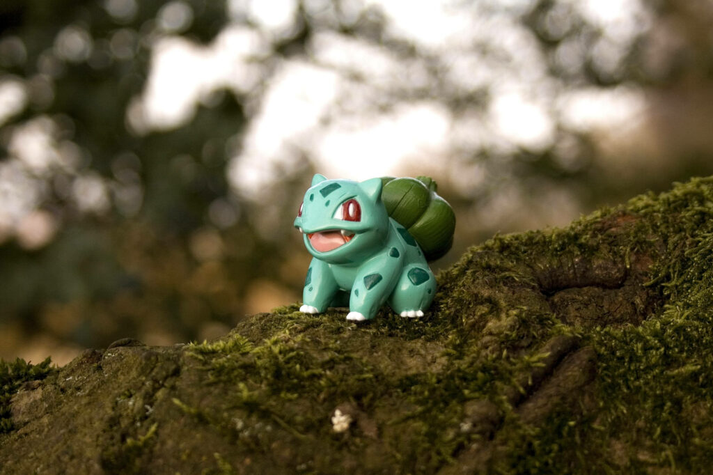 Tiny Bulbasaur toy on mossy tree branch vibrant Pokémon wallpaper background nature blur