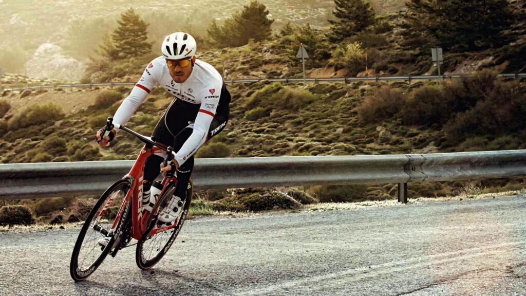 Cyclist in professional gear rides red and white road bike on winding mountain road at sunset Wallpaper