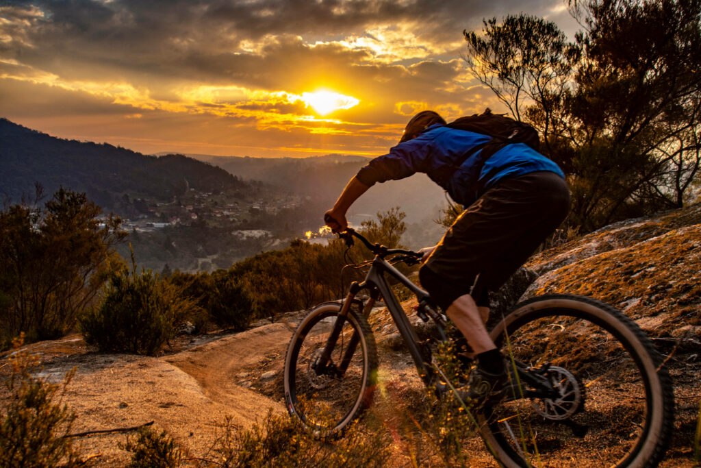 Cyclist biking rugged Tasmanian trail at sunset with rocky terrain and silhouetted trees Wallpaper