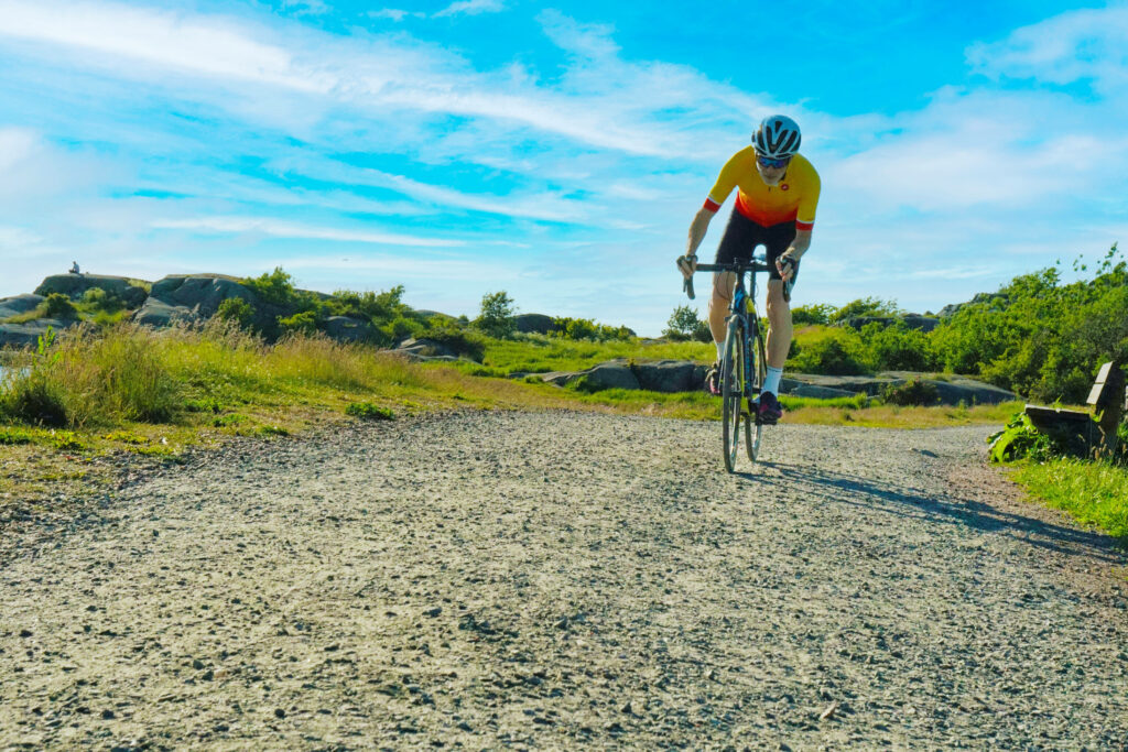 Cyclist in yellow and red jersey rides on sunlit dirt road through green fields and rocky hills Wallpaper