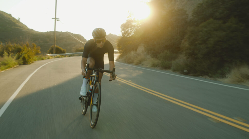 Cyclist in black gear rides fast on sunlit winding road with mountains and trees outdoors Wallpaper